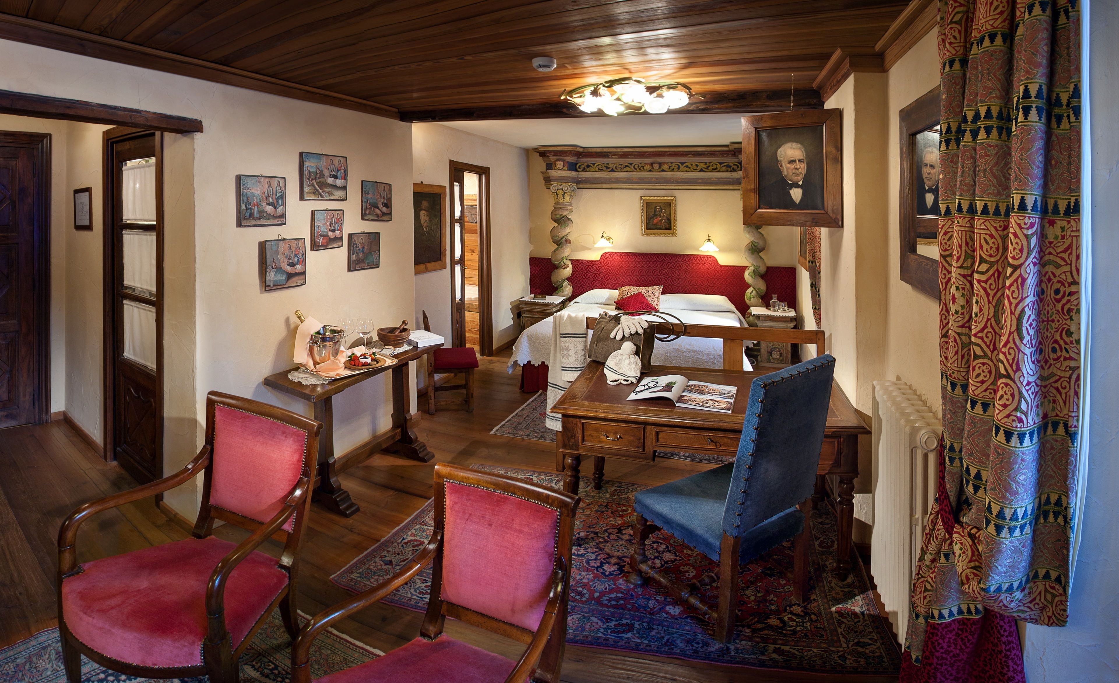 A traditional room with a wooden ceiling, a bed with a red headboard, a desk with a chair, and a dining table with chairs.