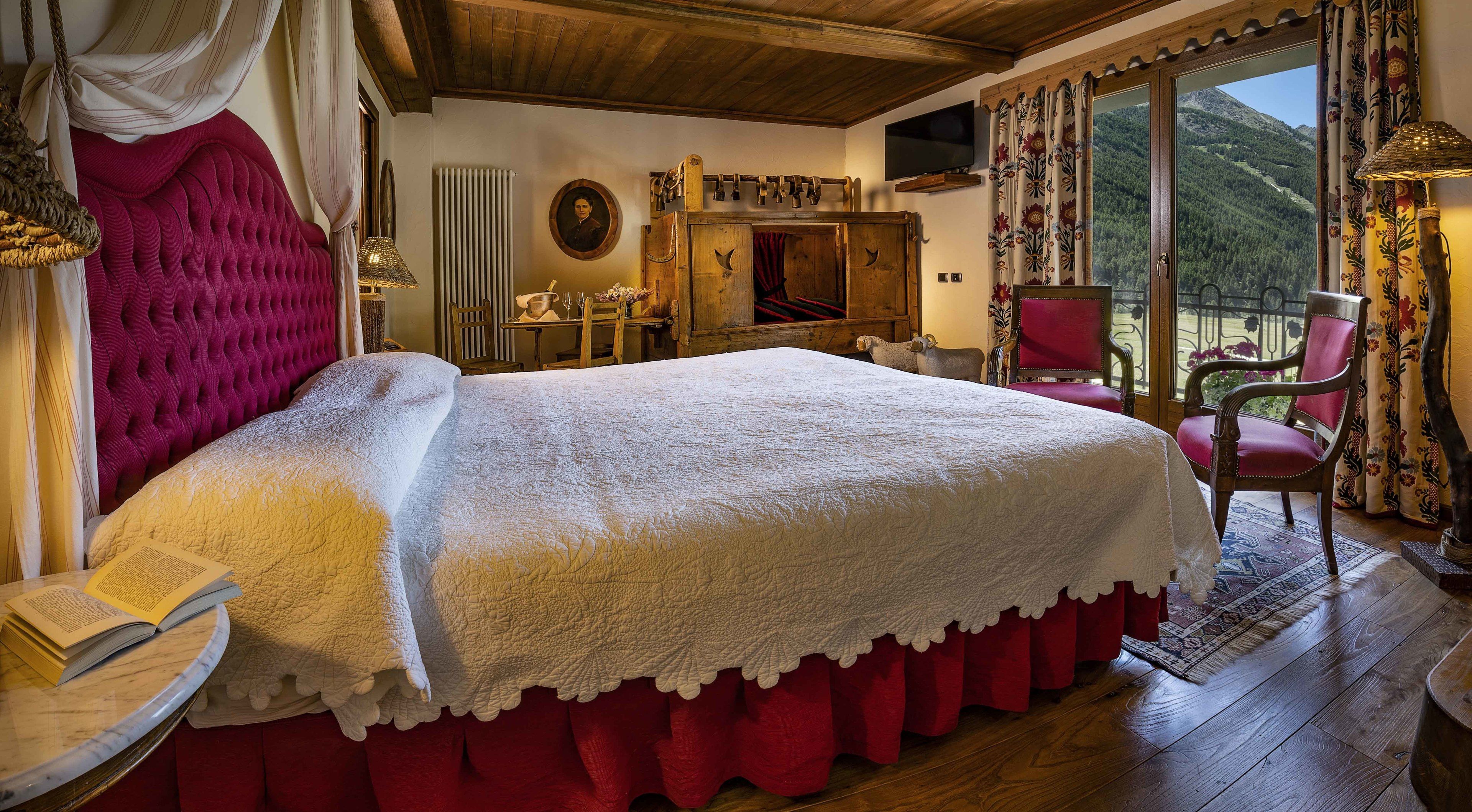 Un dormitorio con una cama grande, cabecera de madera, cómoda de madera, una silla y vistas a un paisaje montañoso a través de una ventana.