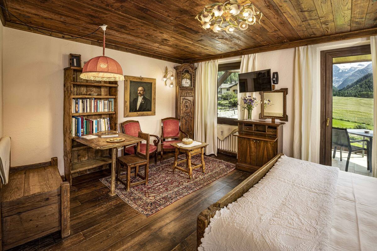 A cozy room with a wooden floor, a bed, a desk with a chair, a bookshelf, a clock, and a view of the outdoors.