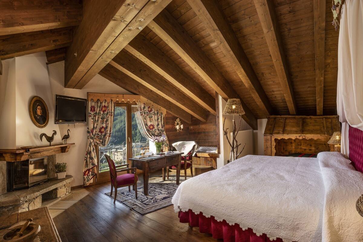 A rustic bedroom with a wooden ceiling, a large bed with a white blanket, a desk with a chair, a fireplace, and a view of the mountains through the window.