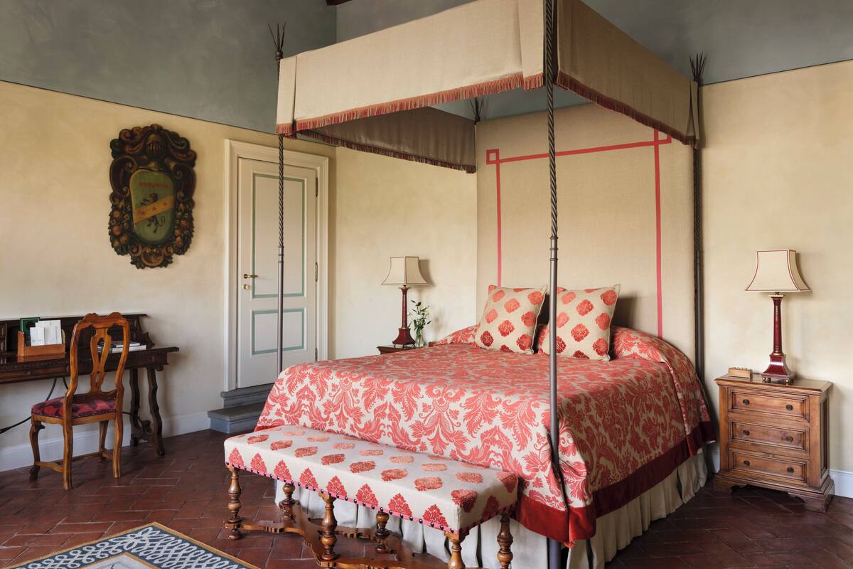 Un lit à baldaquin à quatre poteaux, surmonté d’une couvre-lit à motifs rouges et blancs, une table de chevet en bois avec une lampe, une chaise et un bureau avec une lampe.