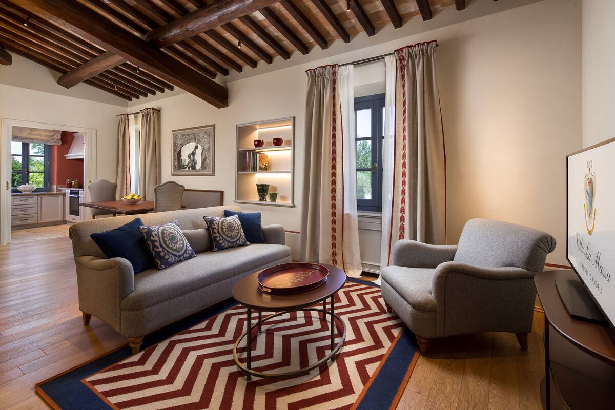 Un salon comprenant un canapé, un fauteuil et une table basse. On aperçoit une cuisine au fond, une fenêtre avec des rideaux et un mur sur lequel se trouve une étagère où sont disposés des objets décoratifs.