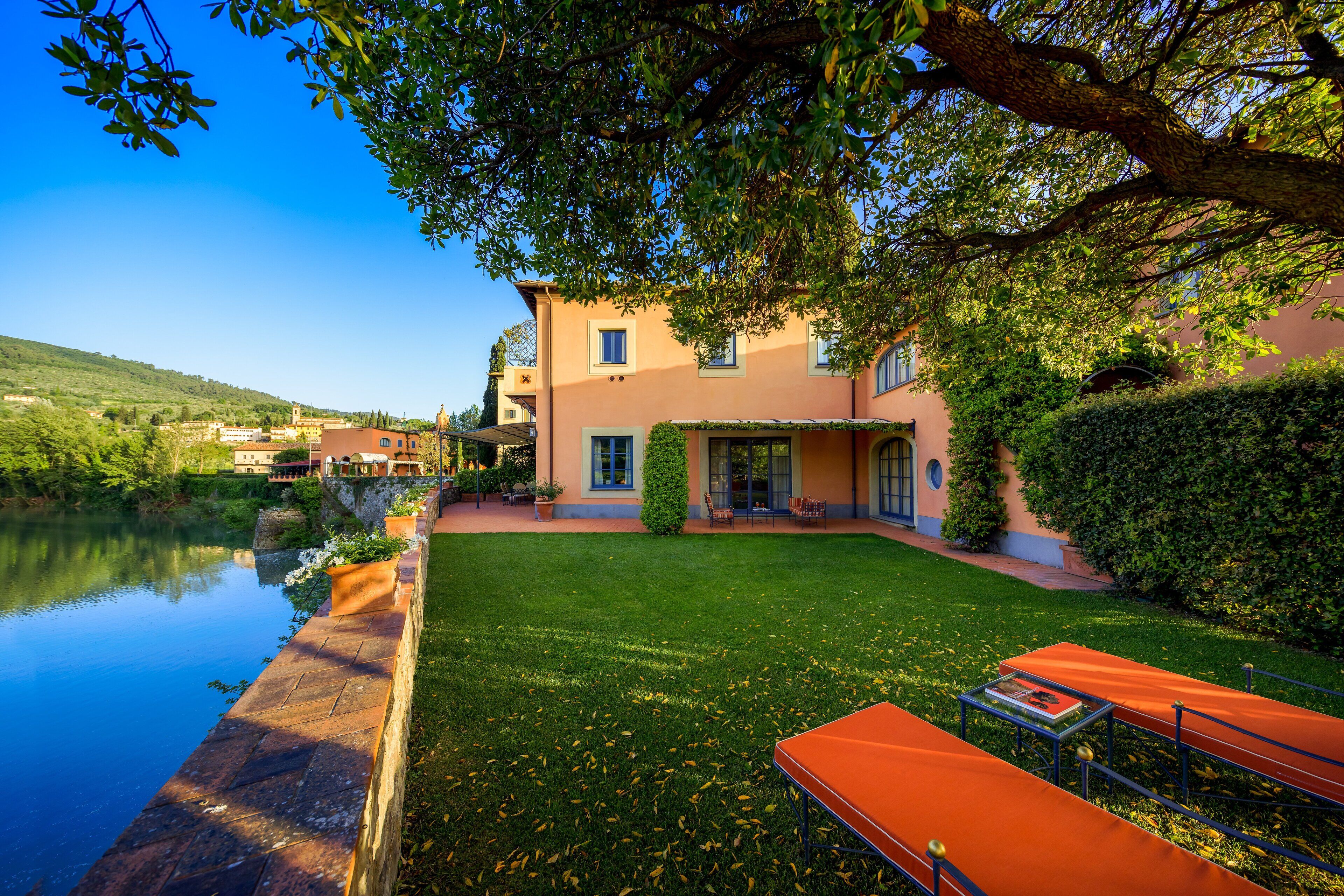 A serene lakeside property with a well-maintained lawn, outdoor seating, and a large tree providing shade.