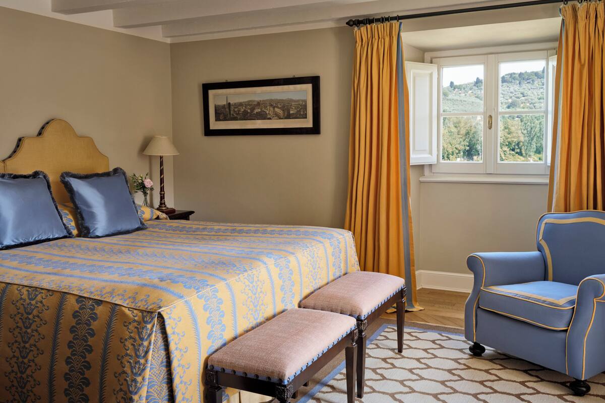 Une chambre à coucher avec un lit, une chaise, un banc, une lampe, un tableau au mur et une fenêtre avec des rideaux.