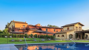 Un área junto a la piscina con sillones y sombrillas, un edificio de piedra y una casa grande de color naranja con tejado de tejas.