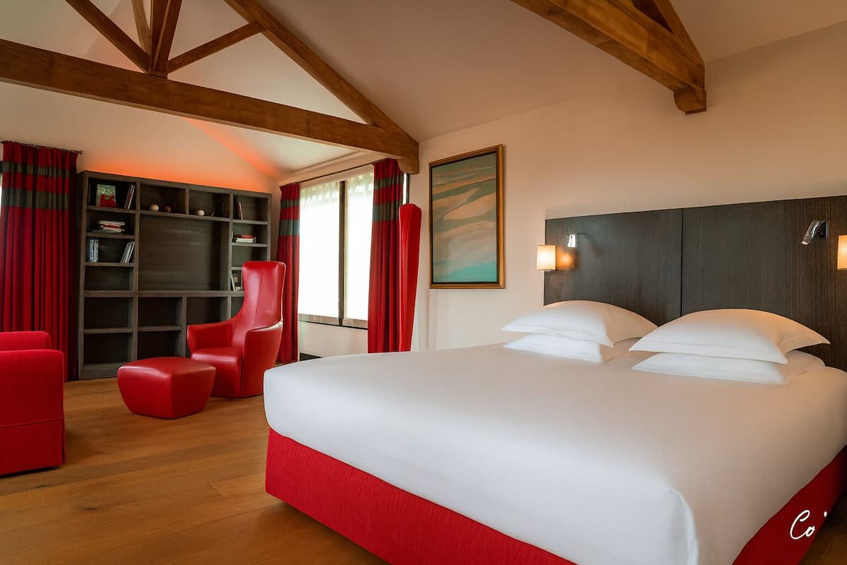 Une chambre d’hôtel avec un grand lit, des fauteuils à revêtement rouge, une bibliothèque et un tableau au mur.