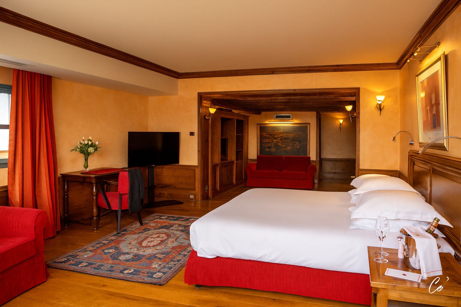 A hotel room with a large bed, a desk with a chair, a television, a red sofa, and a painting on the wall.