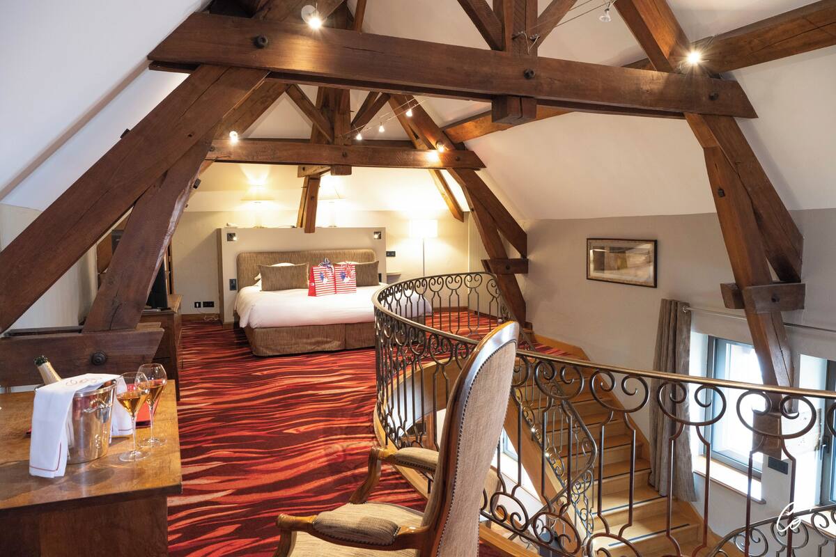 Une chambre avec une moquette rouge, des poutres en bois, un lit, une chaise et une table avec des verres et une bouteille.