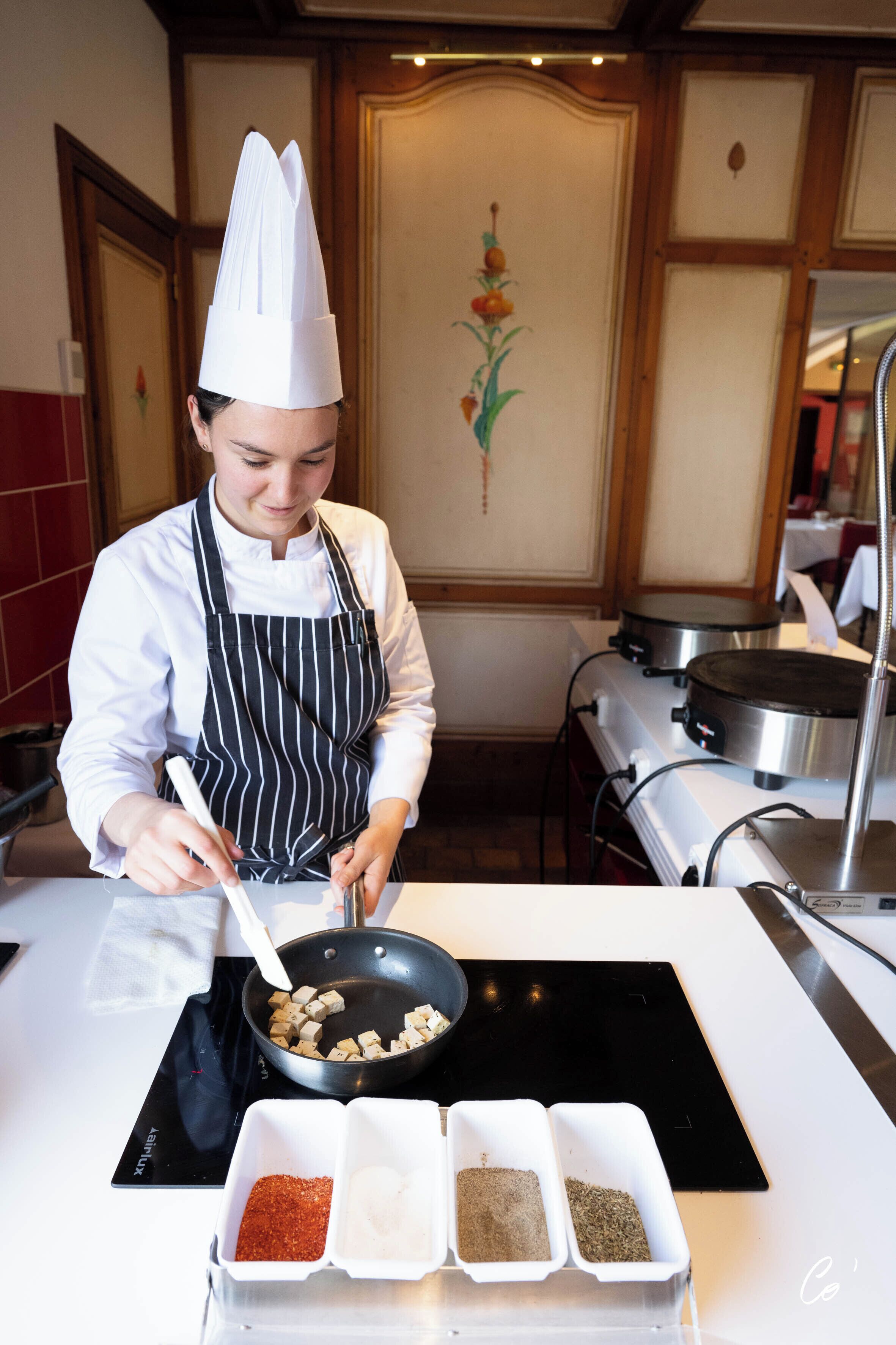 A chef in a kitchen with a stove, a pan, and spice containers.
