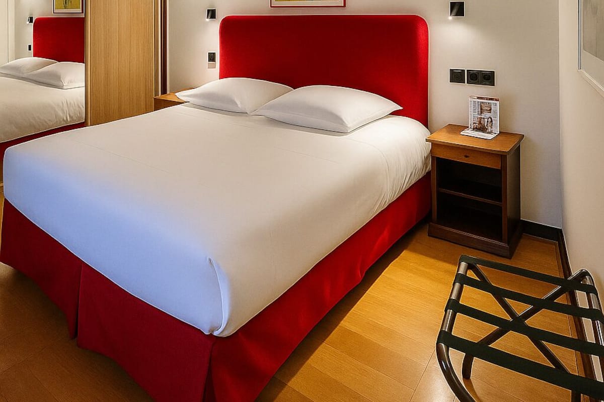 Une chambre d’hôtel avec deux lits, des poutres en bois au plafond, une tête de lit rouge, une table de chevet avec une lampe et un tableau encadré au mur.