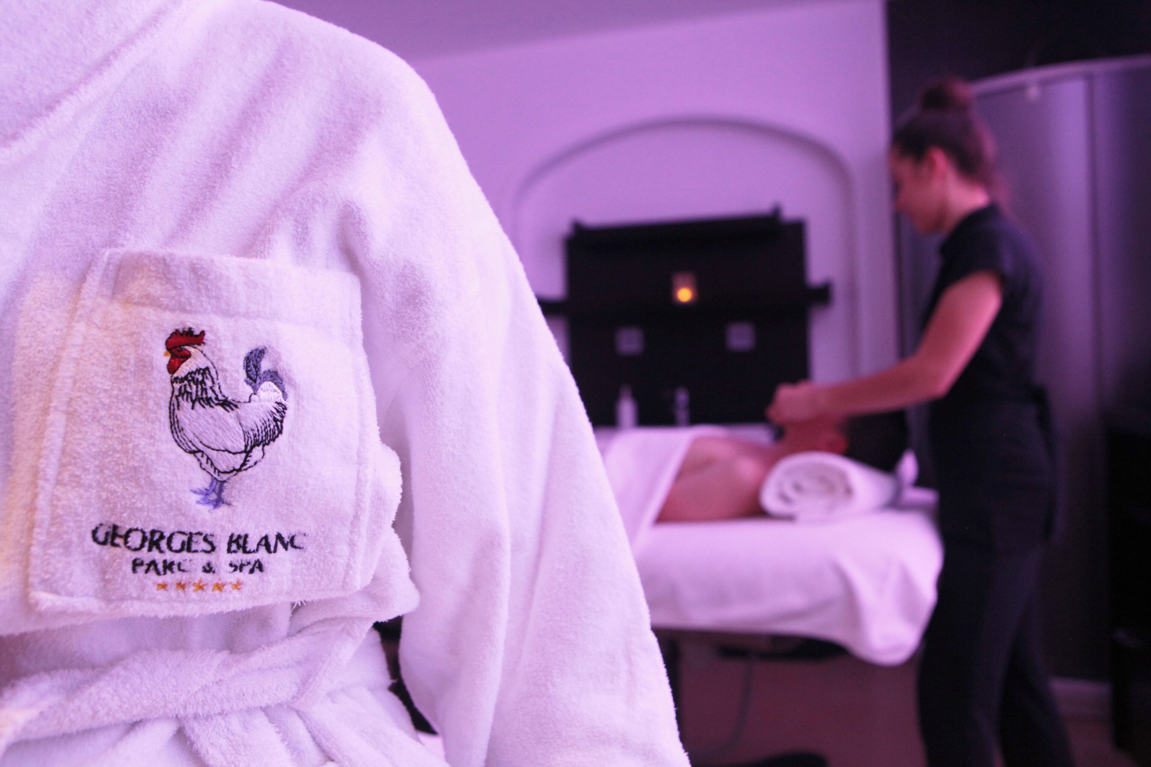 A person in a robe with a rooster logo and text, in a room with a massage table and another person attending to a client.