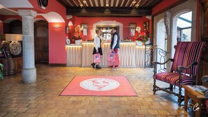 Een receptie met een incheckbalie, een rode loper met een logo van een haan, een houten stoel en een groot raam.