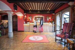 Un área de recepción con mostrador de check-in, una alfombra roja con el logotipo de un gallo, una silla de madera y un ventanal grande.