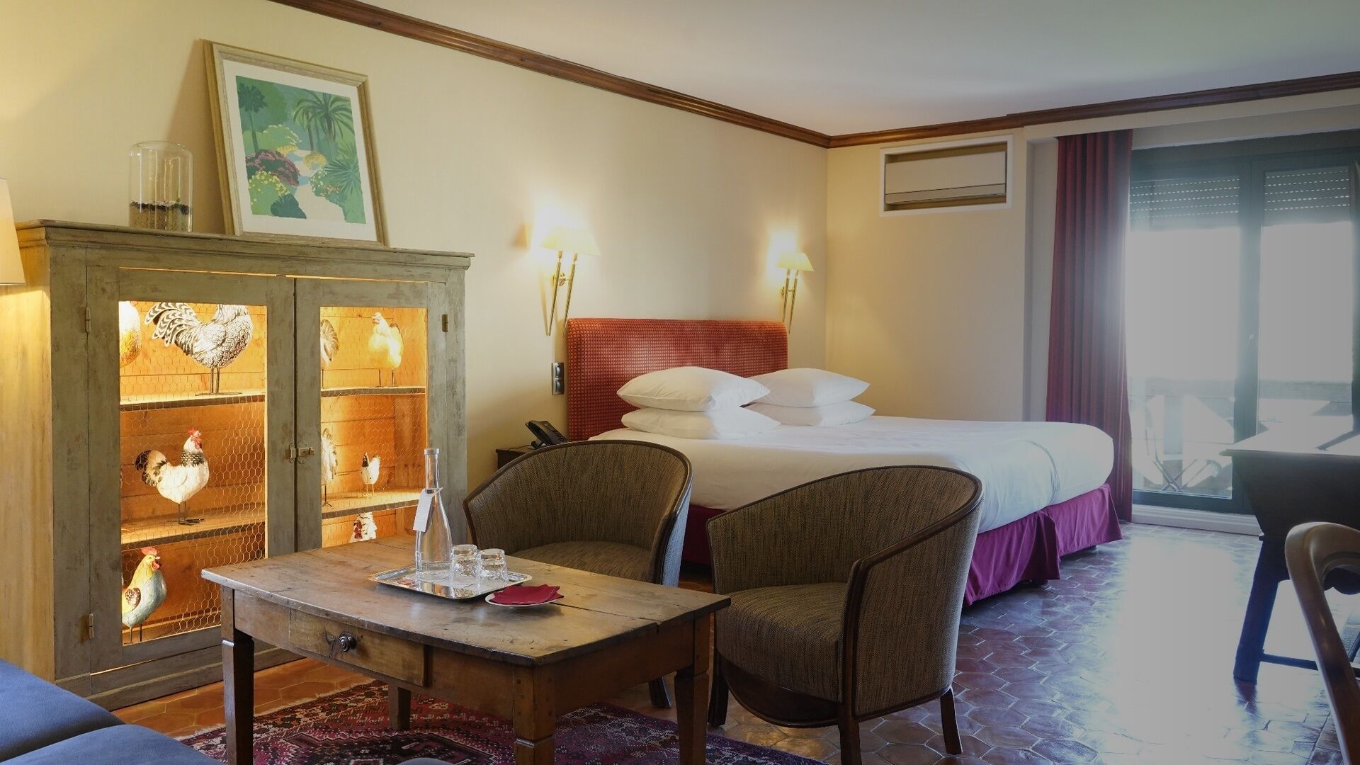 Habitación de hotel con cama, aparador de madera con puertas de cristal, mesa con sillas y vista al exterior por una ventana.