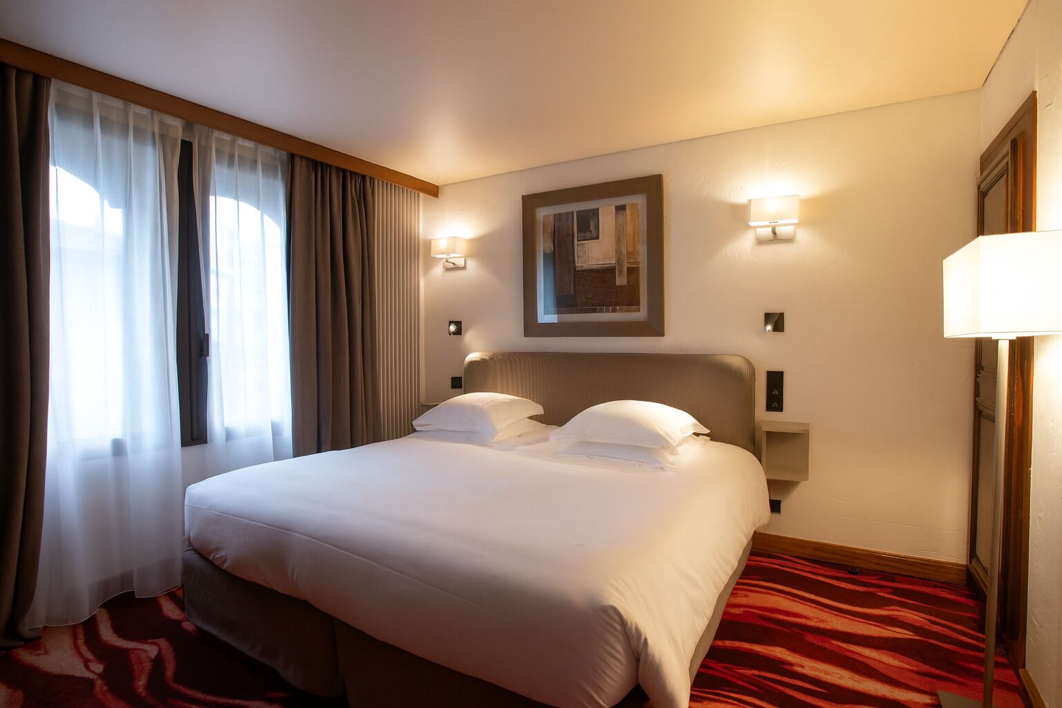 A hotel room with a large bed, two bedside lamps, a painting on the wall, and a red carpet.