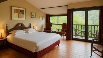 A hotel room with a large bed, a desk with a chair, a balcony with outdoor seating, and a view of greenery.