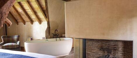 A bedroom with a large bed, a bathtub, a plaid armchair, and a fireplace.