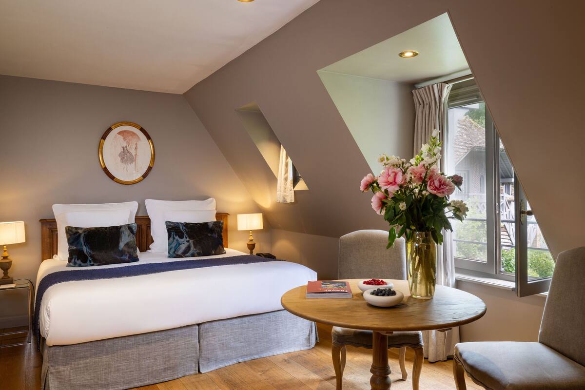 A hotel room with a large bed, a bedside table with a book and a vase of flowers, a chair, and a window with curtains.