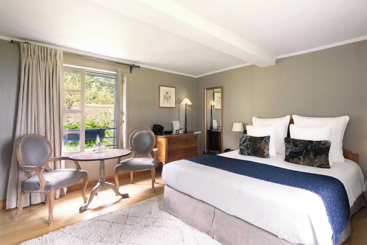 A bedroom with a bed, two chairs, a small table, a dresser, a mirror, and a window with curtains.