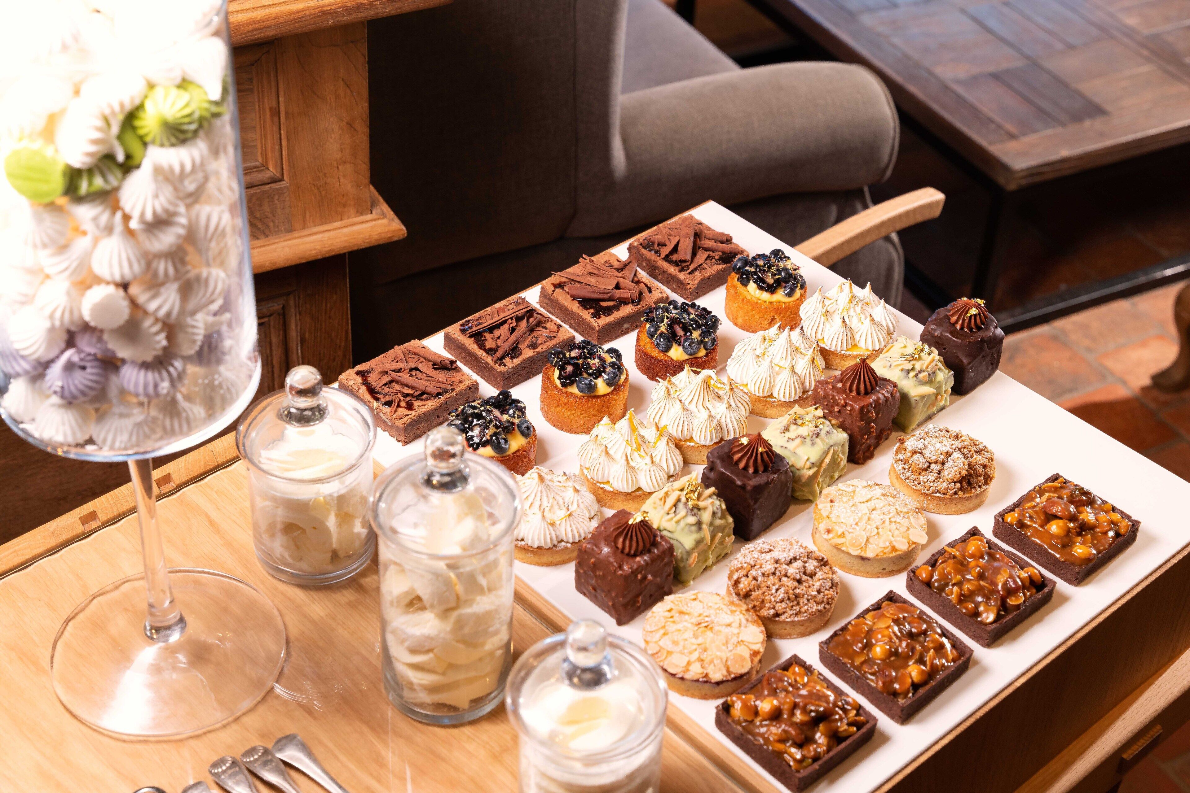 Una variedad de postres en una bandeja, incluyendo cupcakes, brownies y pastelería, con un tarro de crema y una copa de vino.