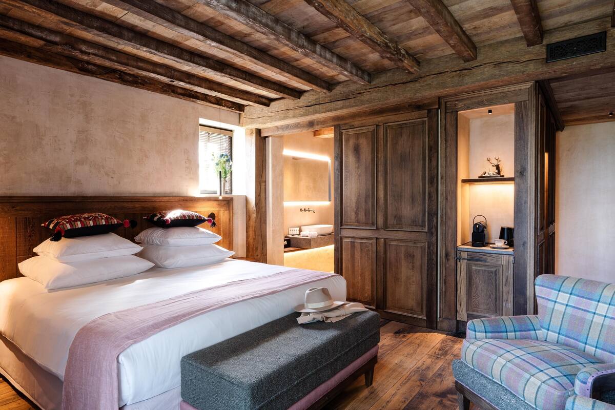 A bedroom with a wooden ceiling, a large bed with multiple pillows, a bench, and a plaid armchair.
