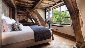 Une chambre avec un plafond en bois, une grande fenêtre donnant sur un paysage verdoyant, un lit avec des oreillers et une table de chevet ornée d’un vase de fleurs.