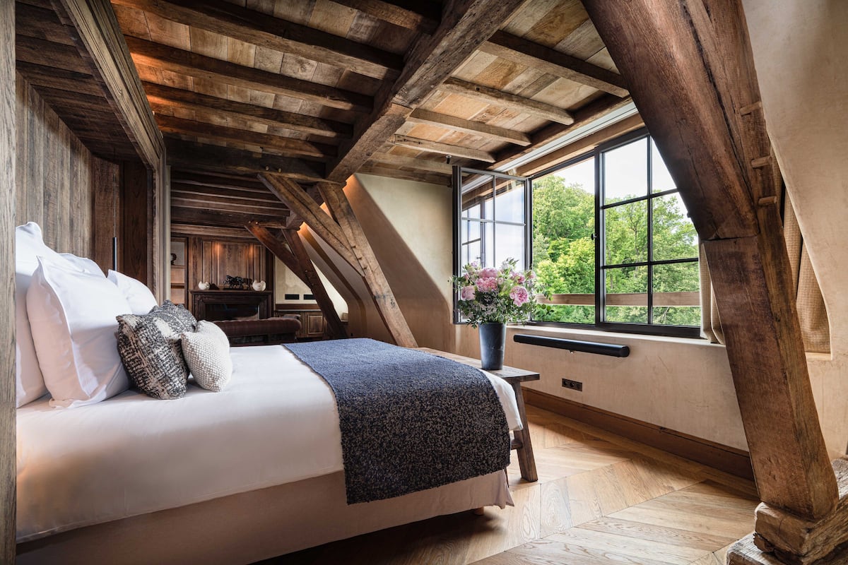 A bedroom with a wooden ceiling, a large window with a view of greenery, a bed with pillows, and a bedside table with a vase of flowers.