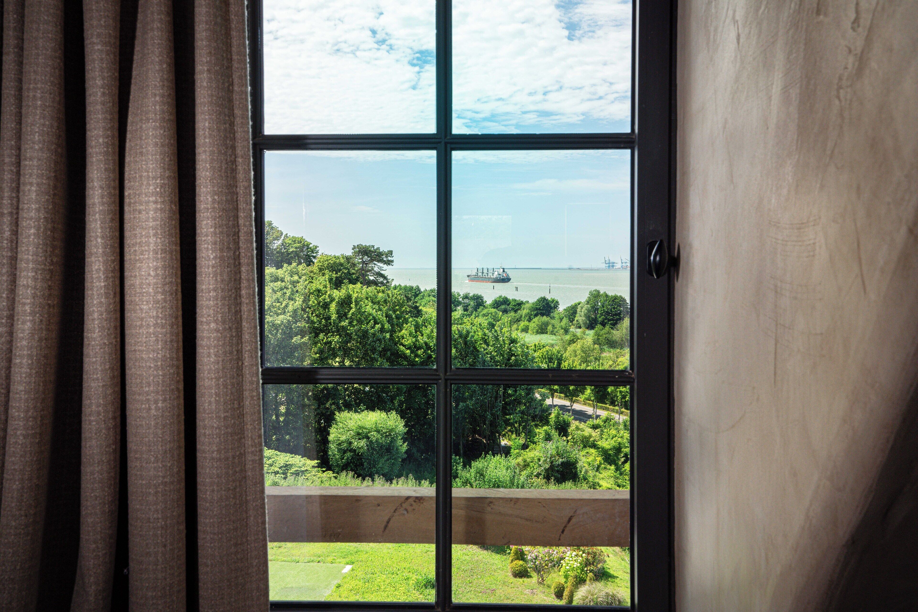 A window with a view of a green landscape and a distant ship.
