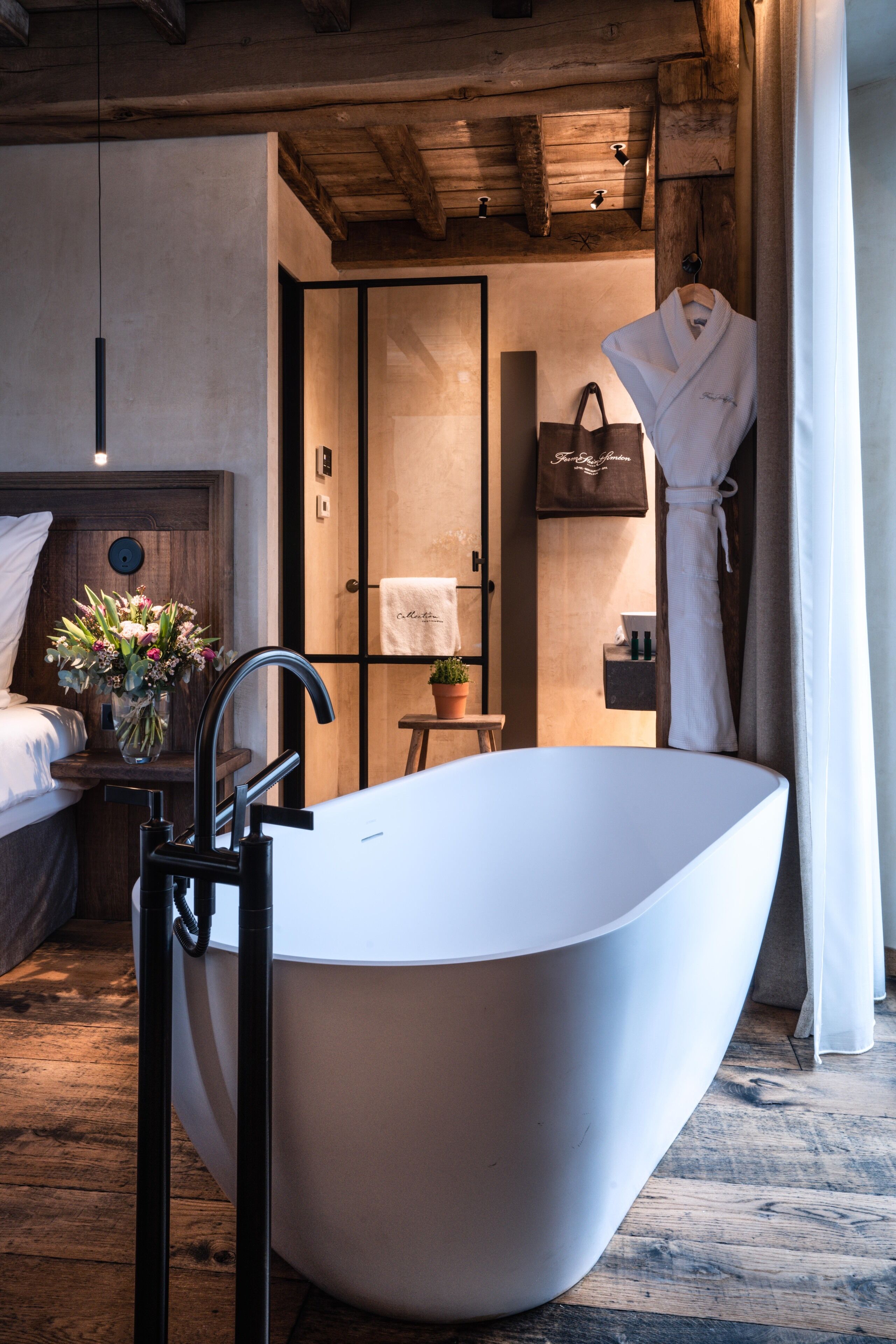 A modern bathroom with a freestanding bathtub, wooden ceiling beams, a towel rack, and a potted plant.