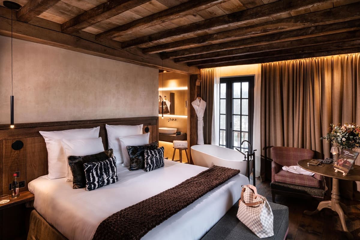 A hotel room with a wooden ceiling, a large bed with white bedding, a sitting area with a chair and table, a bathroom with a bathtub, and a window with curtains.