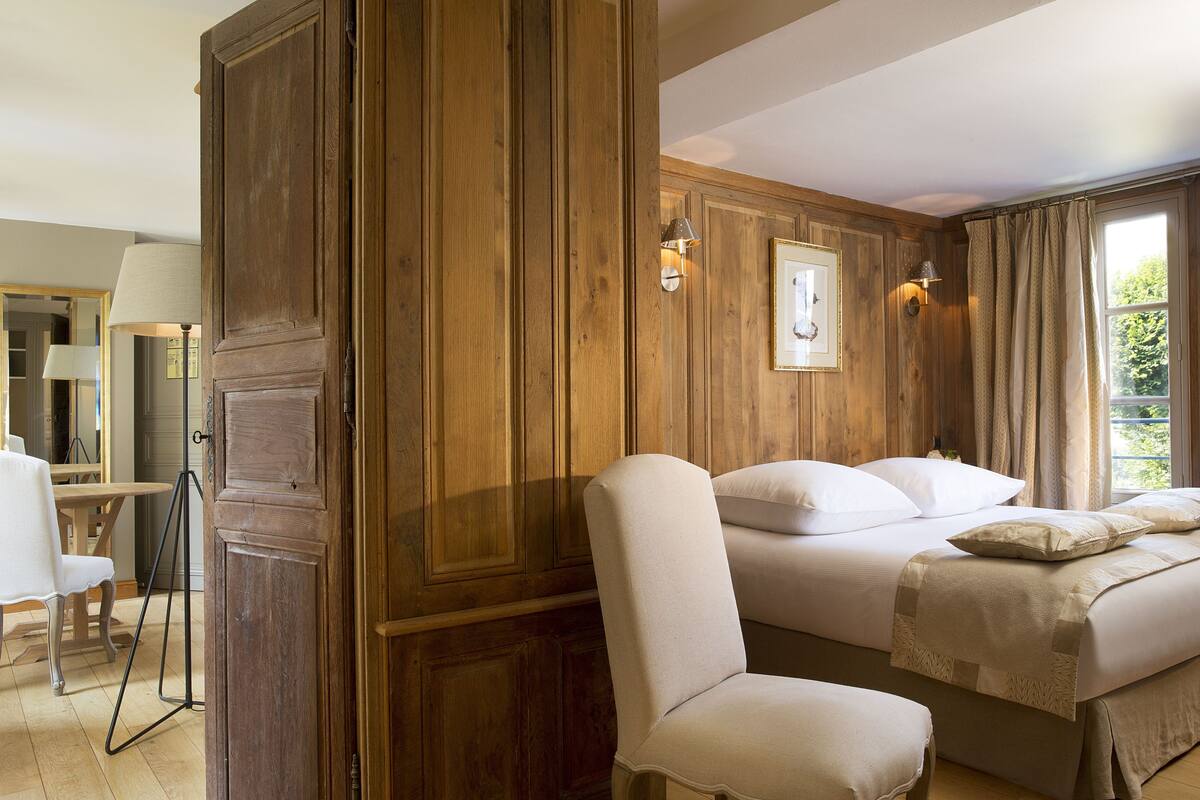 A room with wooden paneling, a bed with white linens, a chair, and a desk with a lamp.