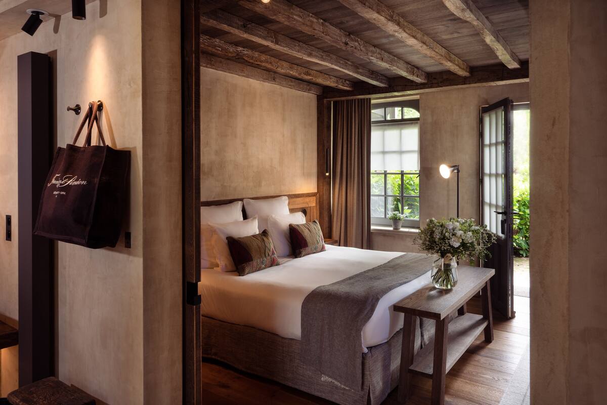 A bedroom with a wooden bed, a bedside table, a nightstand, a large window with curtains, and a wall-mounted bag rack.