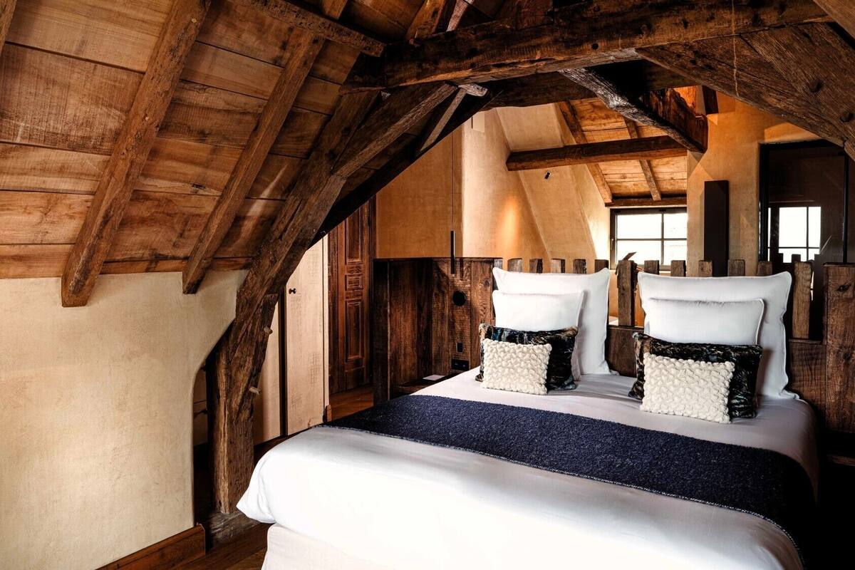 A rustic bedroom with exposed wooden beams, a large bed with white linens and textured pillows, and a view of the outside through a window.