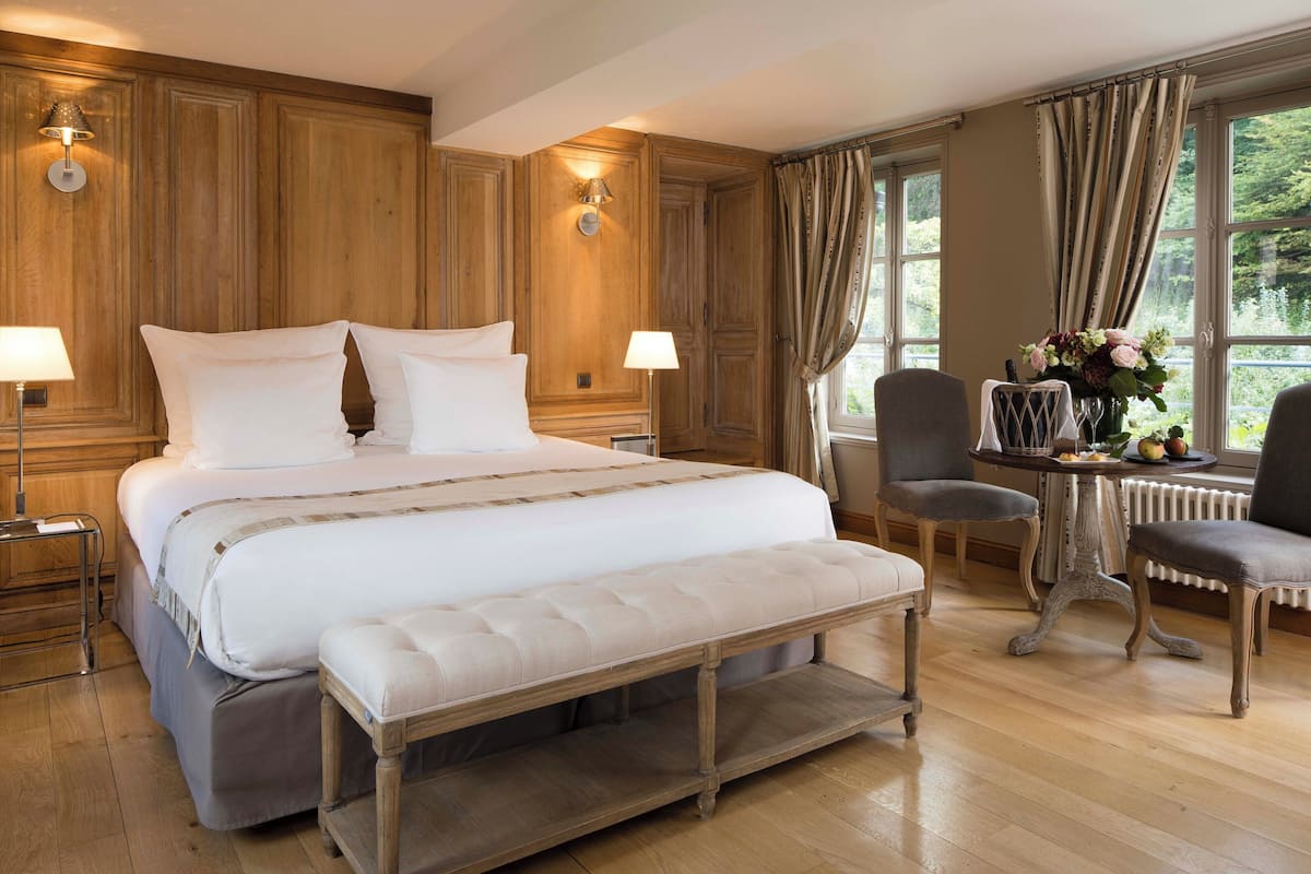 A hotel room with a large bed, two bedside lamps, a bench, a small table with a vase of flowers, and a view of greenery through the window.