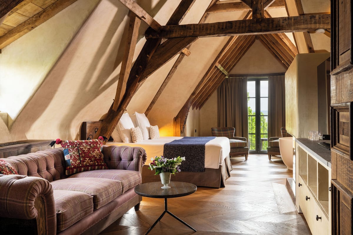 A cozy attic bedroom with a bed, two sofas, a small table with a vase, wooden beams, and a large window.