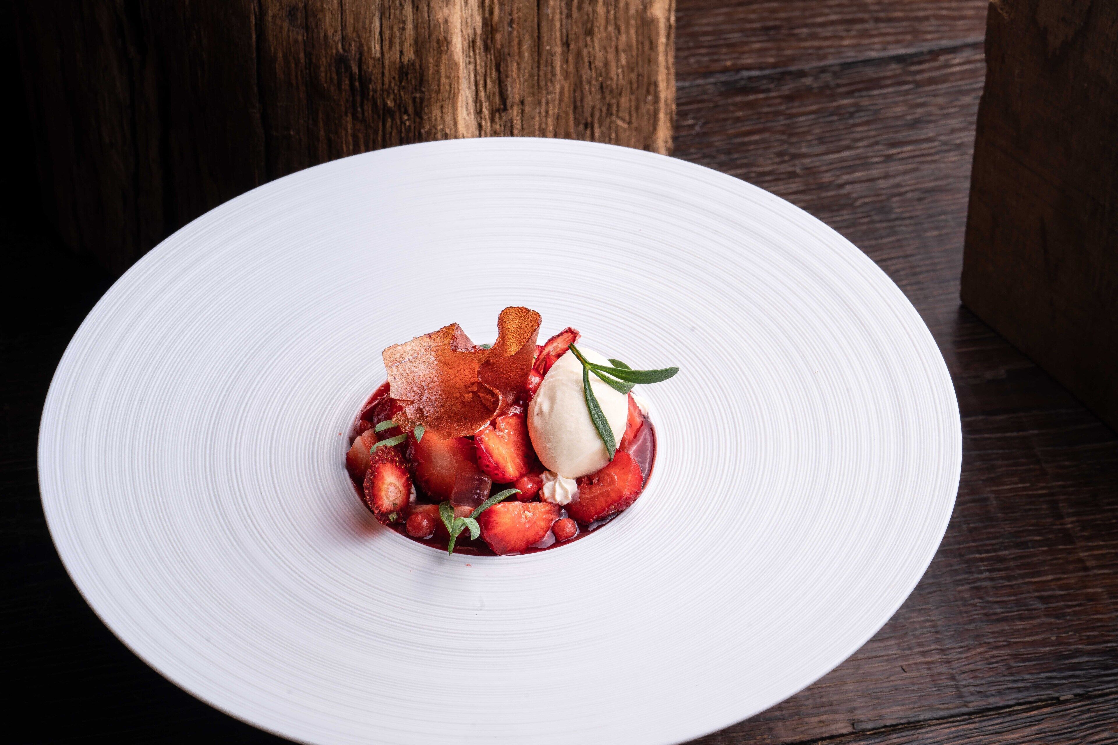 A white plate with a dessert consisting of strawberries, a scoop of ice cream, and a garnish of herbs.