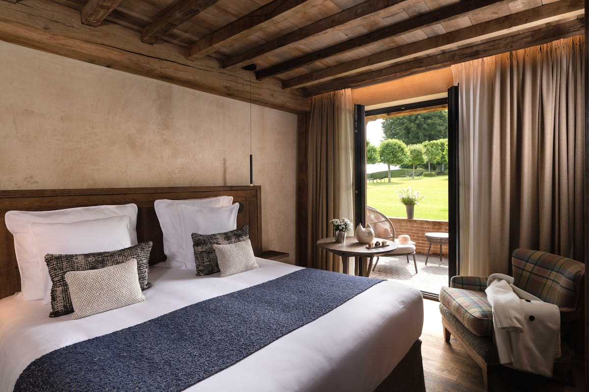 A hotel room with a large bed, a wooden ceiling, a view of a garden through a window, and a small table with a chair.