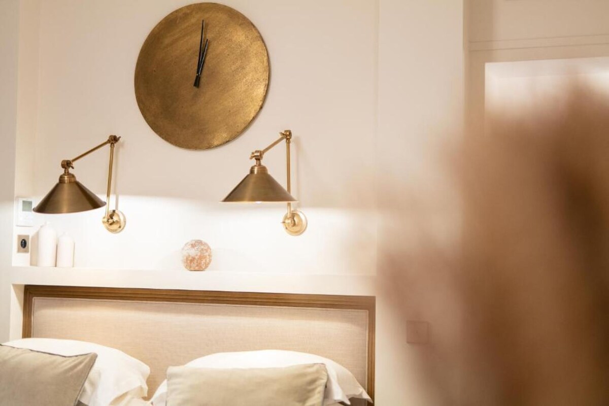 A hotel room with a round wall clock, two bedside lamps, a bed with pillows, and a small decorative item on the nightstand.