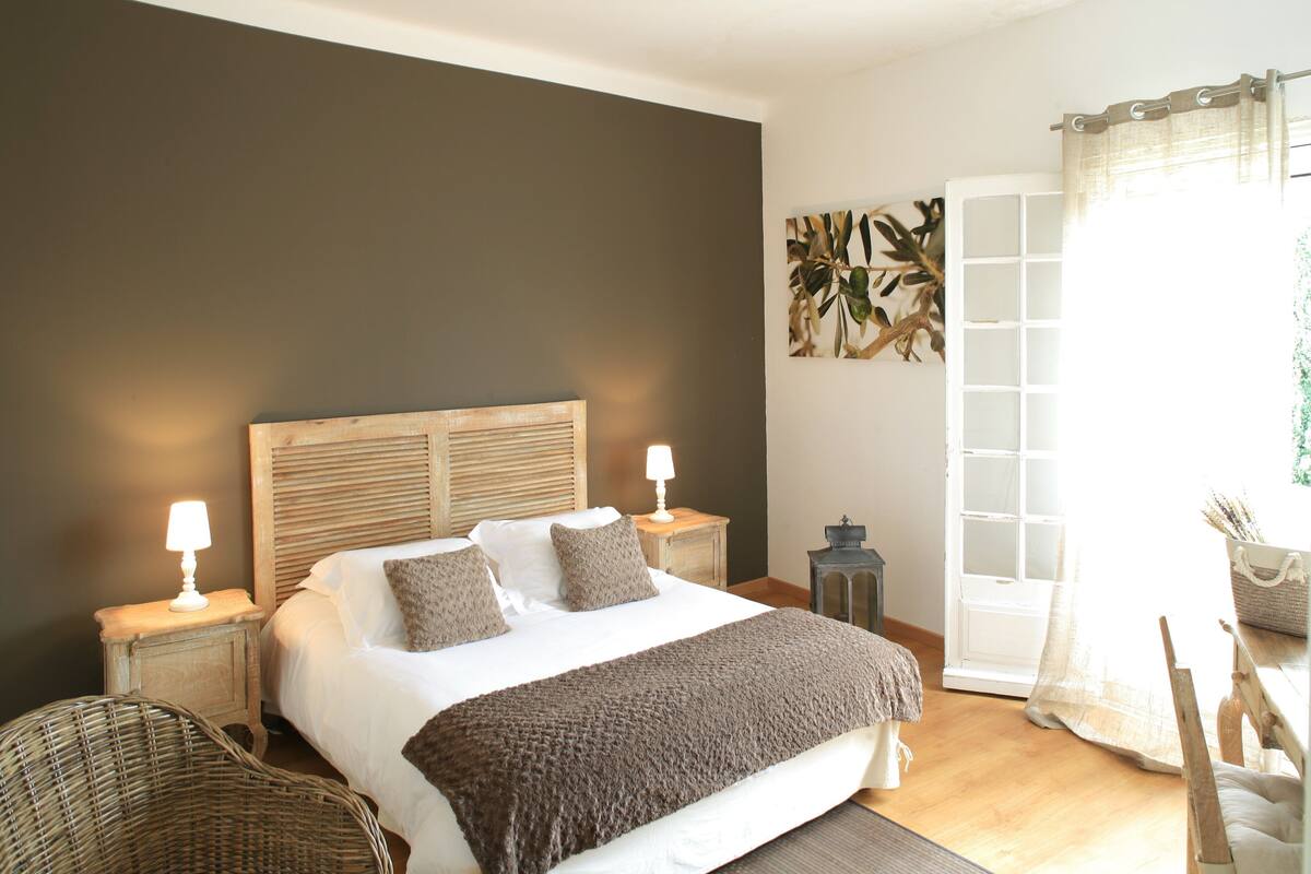 A bedroom with a large bed, two bedside tables with lamps, a wicker chair, a wooden headboard, and a view of the outdoors through a door with curtains.
