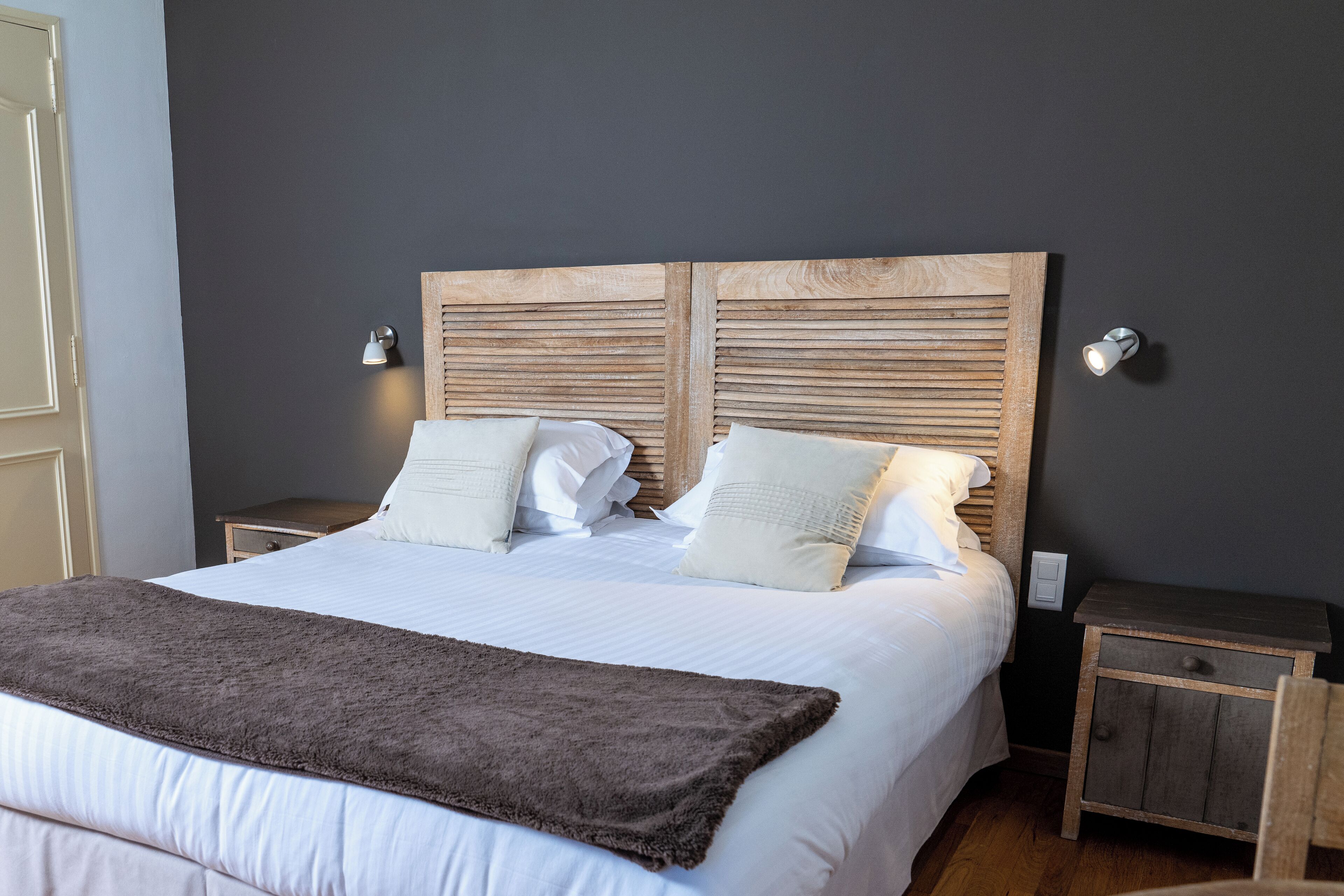 Een netjes opgemaakt bed met een houten hoofdbord, wit beddengoed en een bruine plaid. Er staan twee nachtkastjes met lampen bij, en een klein houten kastje.