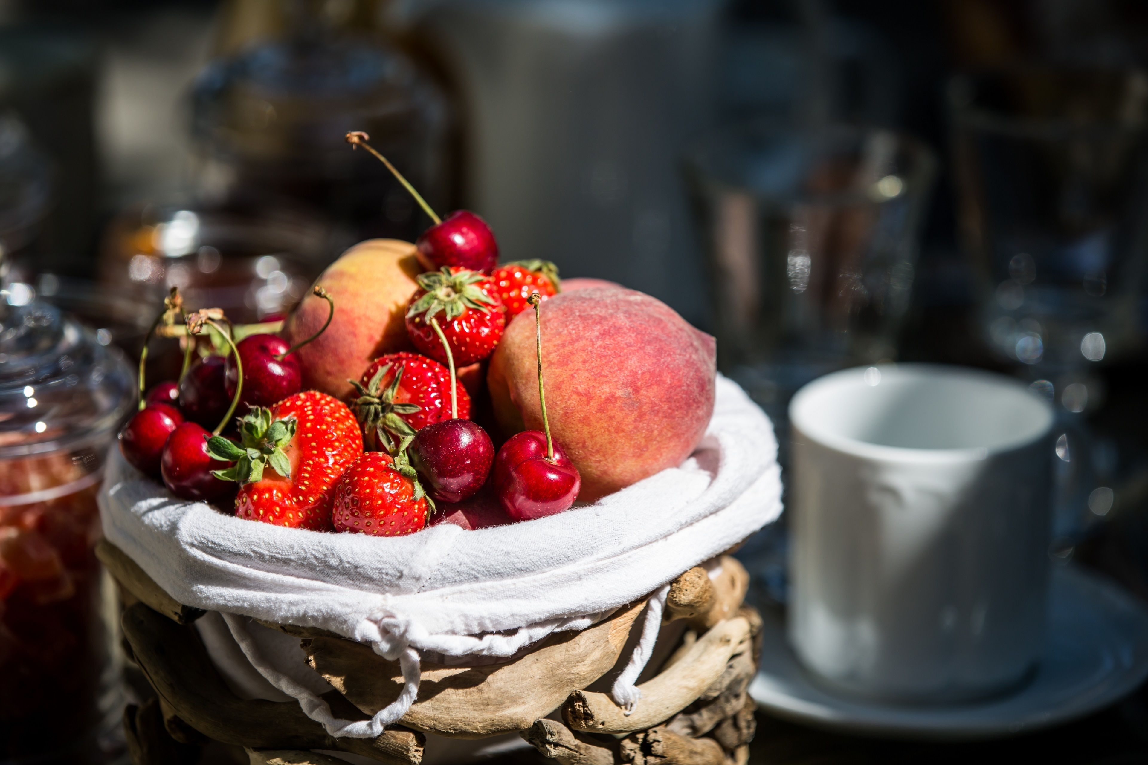 Un panier rempli de fruits frais, notamment des fraises, des cerises et des pêches, posé sur une surface tressée recouverte d’un tissu blanc.