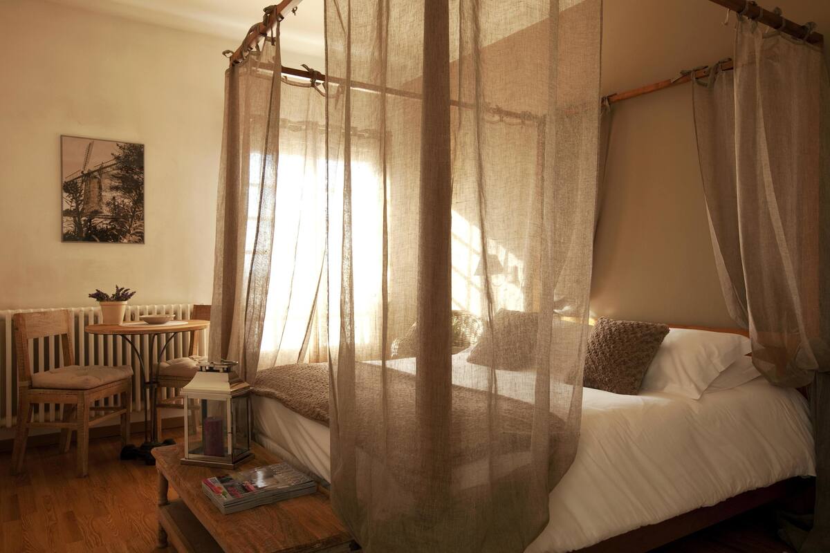 A bedroom with a canopy bed, a small table with a plant, a chair, a bench, and a wall-mounted picture.