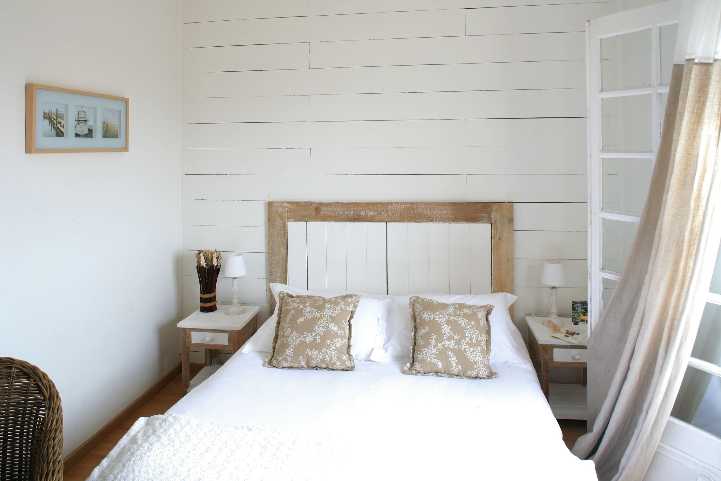 A bedroom with a wooden headboard, two bedside tables with lamps, a wicker chair, and a framed picture on the wall.