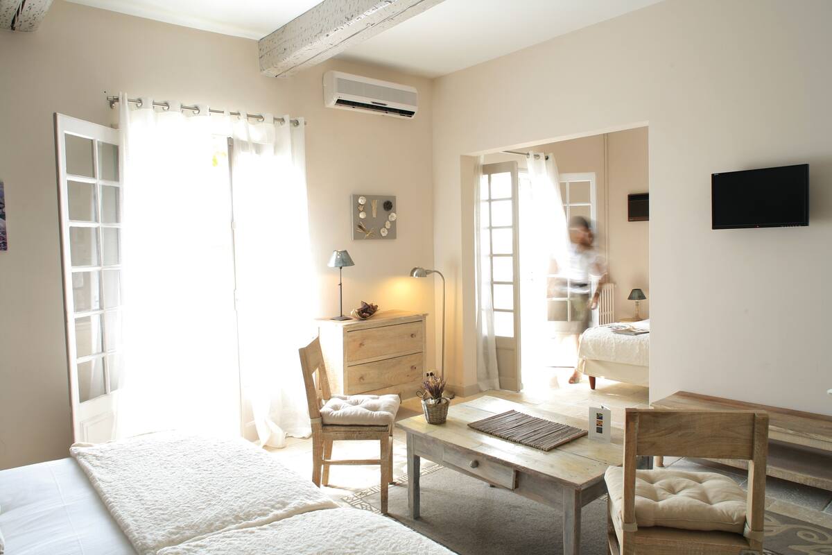 A bright, well-lit room with a bed, a desk, and a chair. There is a window with curtains, a wall-mounted TV, and an air conditioning unit.