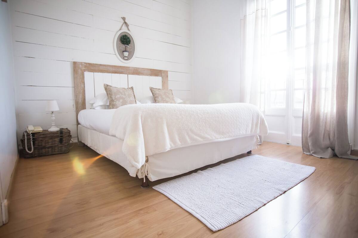 A bedroom with a wooden headboard, a white bedspread, a bedside lamp, and a wicker basket.