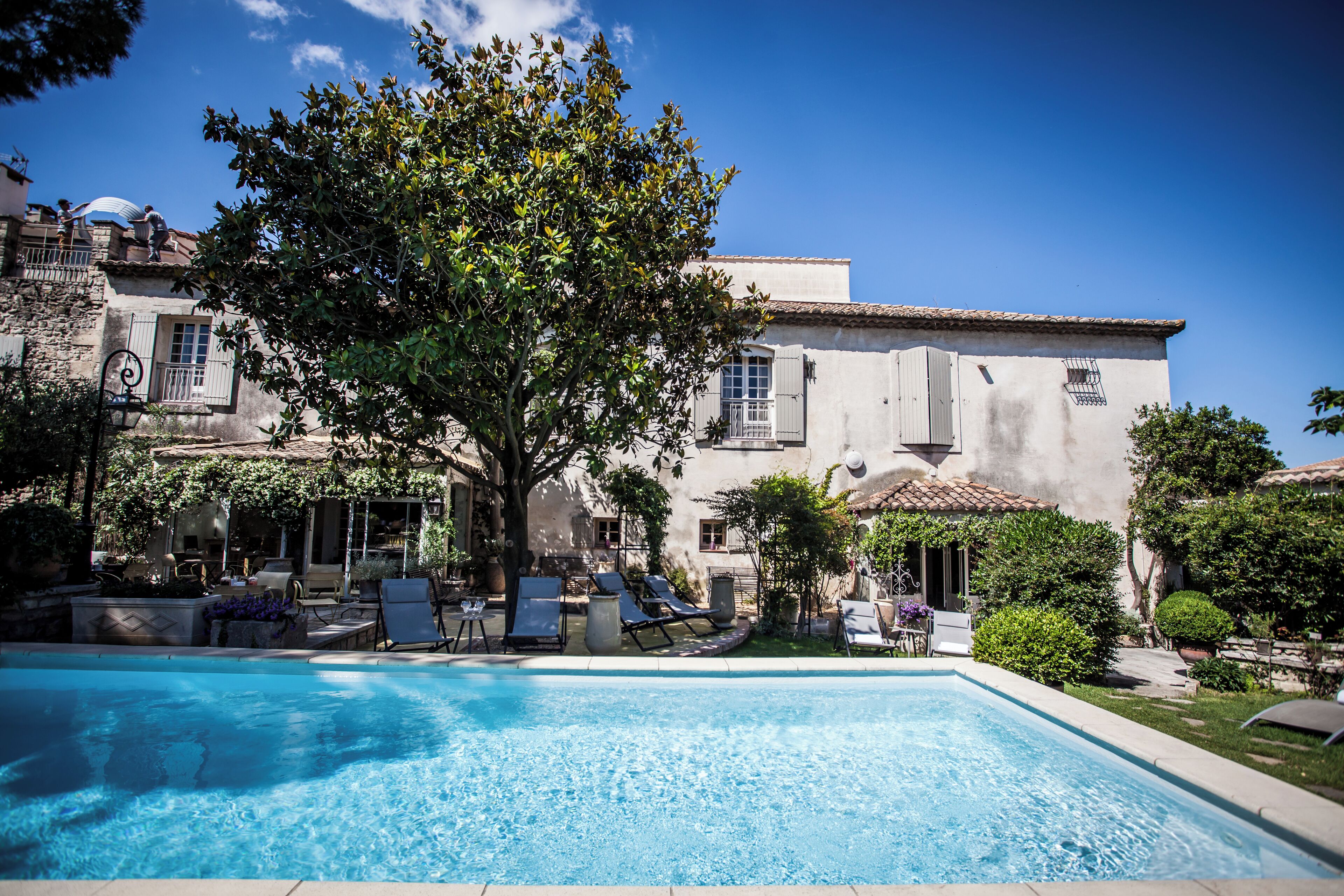 A swimming pool with lounge chairs and a tree in the foreground, surrounded by a building with a tiled roof and windows.