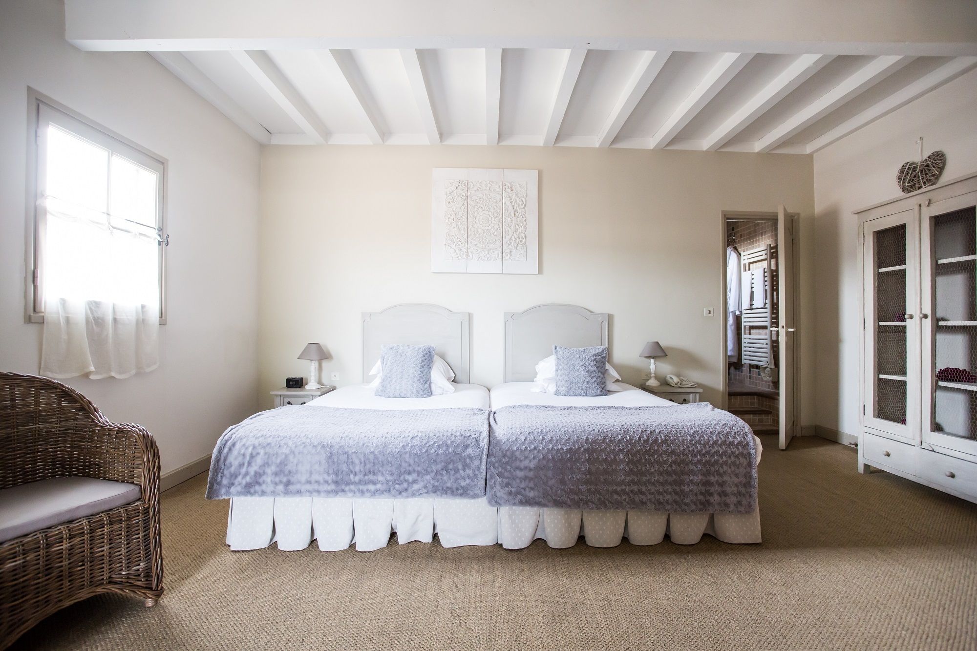 A double bed with a white headboard and a matching footboard, a wicker chair, a bedside table with a lamp, and a wardrobe with a mirror.