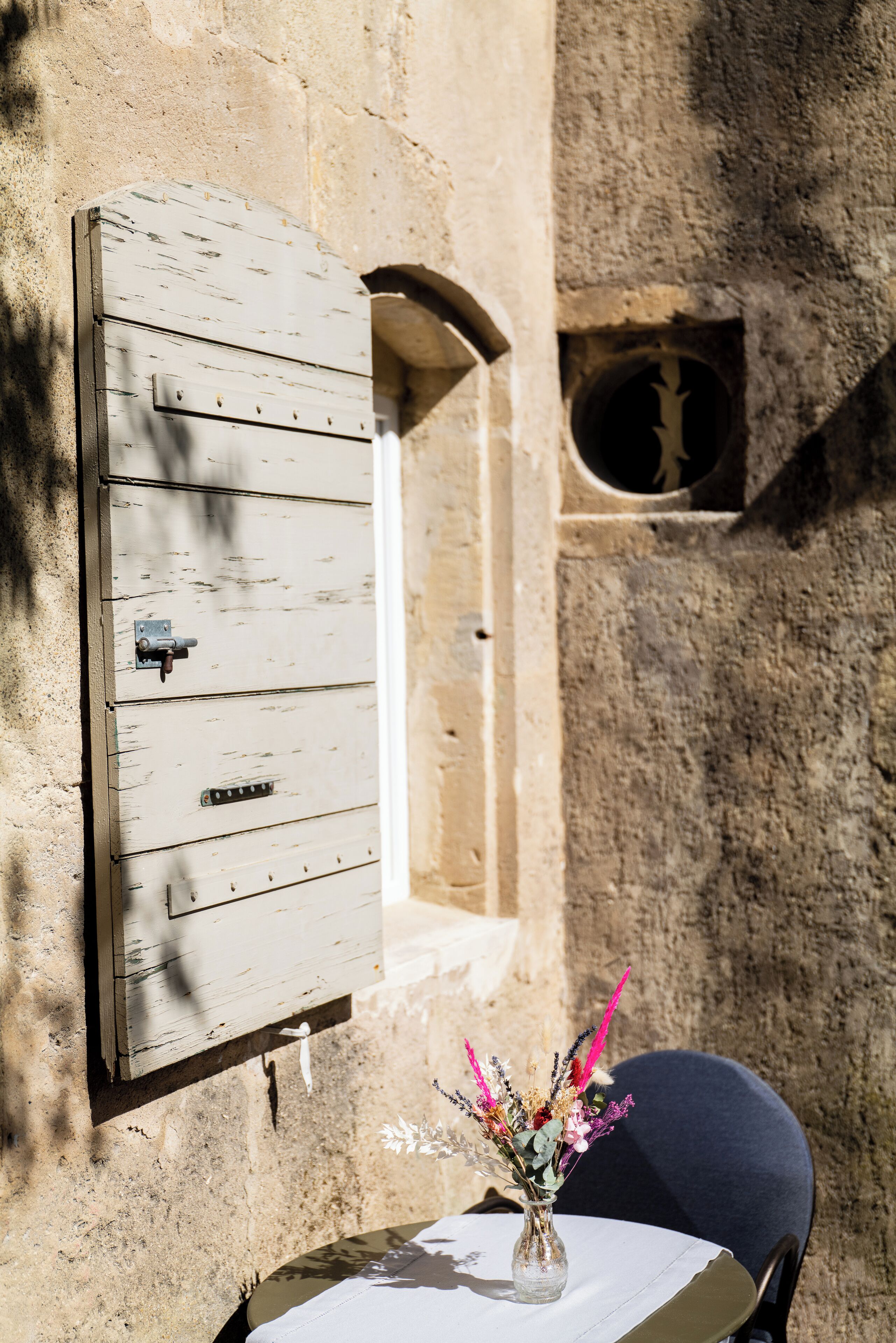 A rustic stone wall with a wooden shutter and a small window. A table with a vase of flowers is visible in the foreground.