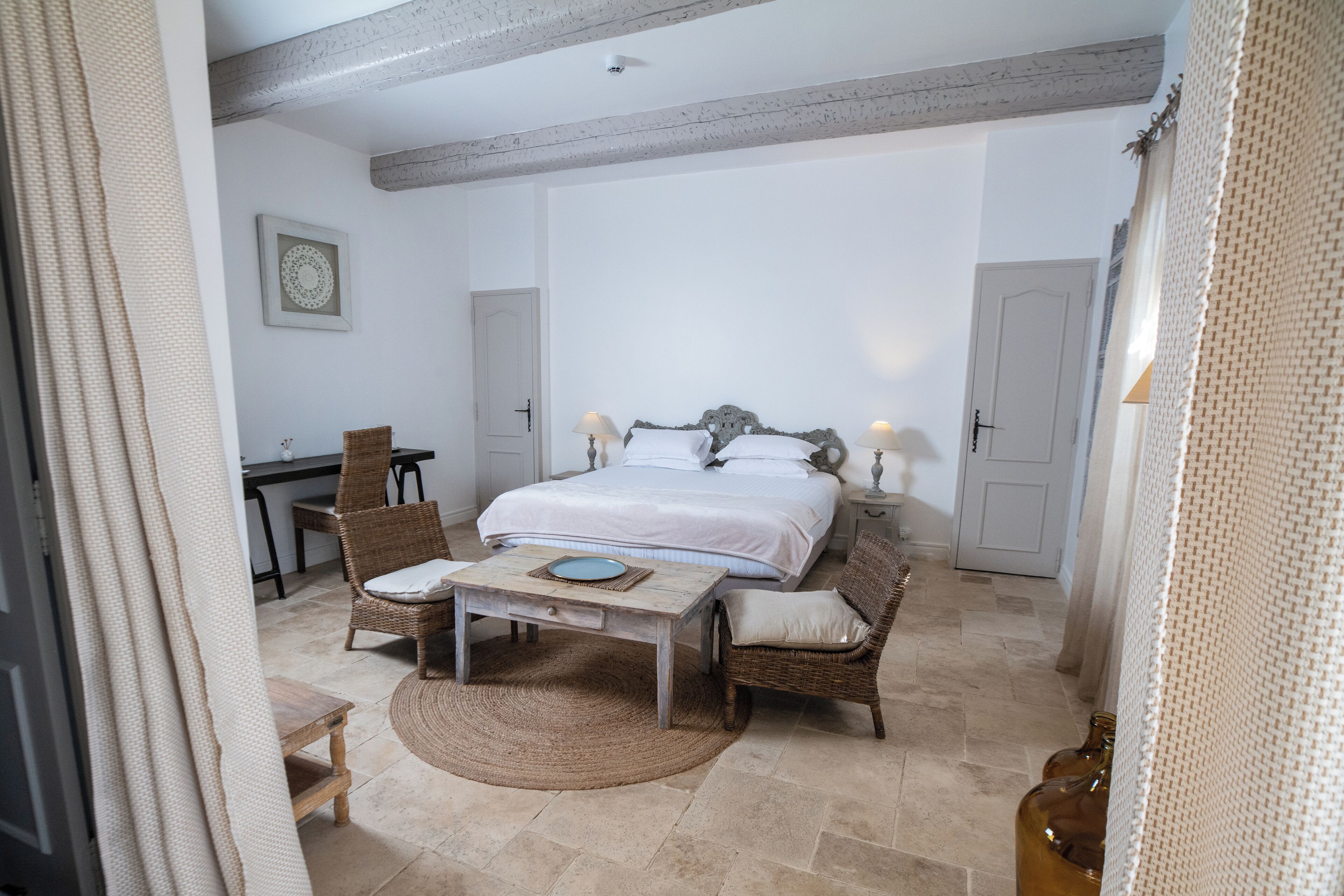 Een slaapkamer met een groot bed, twee rieten stoelen, een tafeltje en een deur.