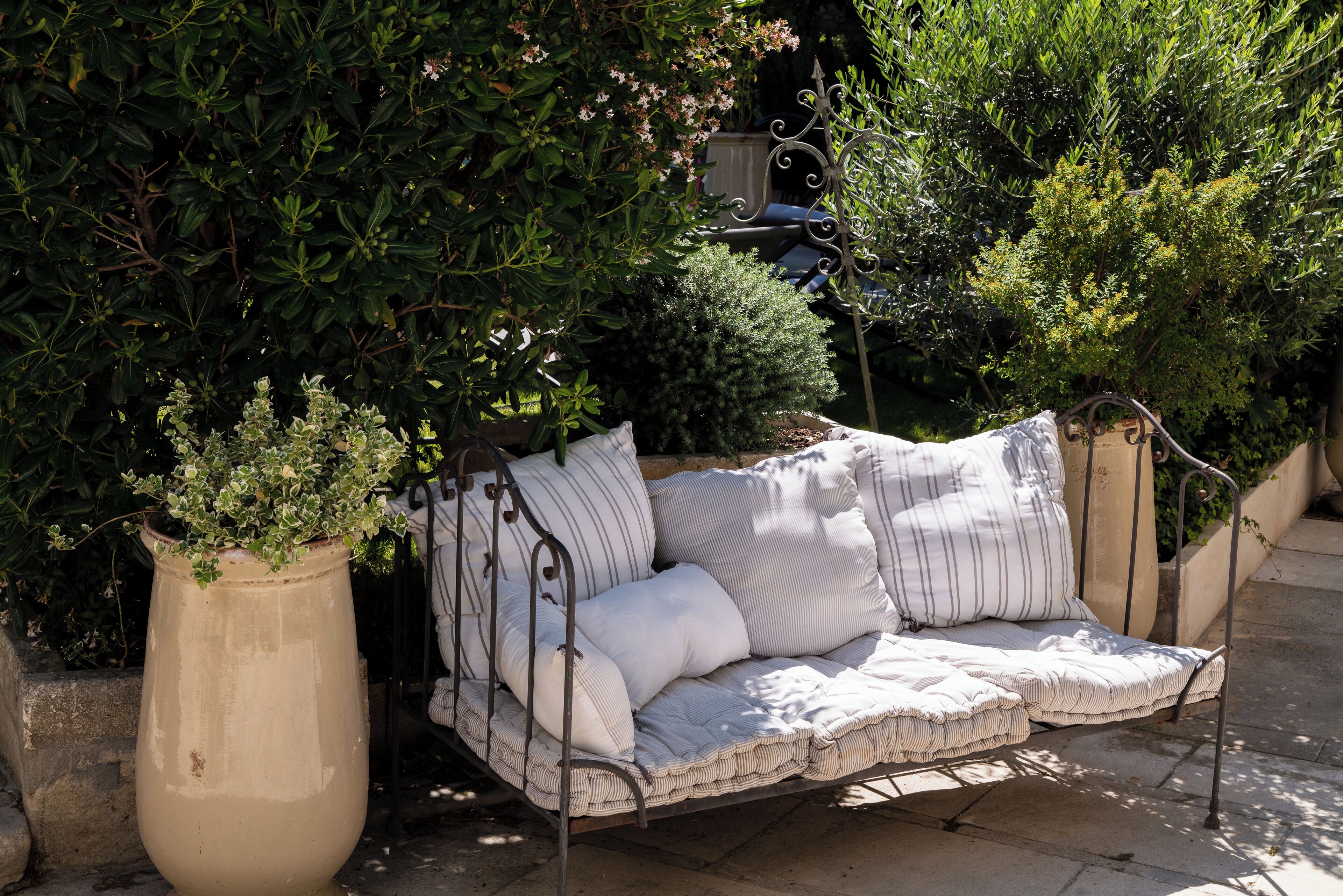 A wrought iron outdoor sofa with striped cushions, positioned next to a large planter with a small tree.